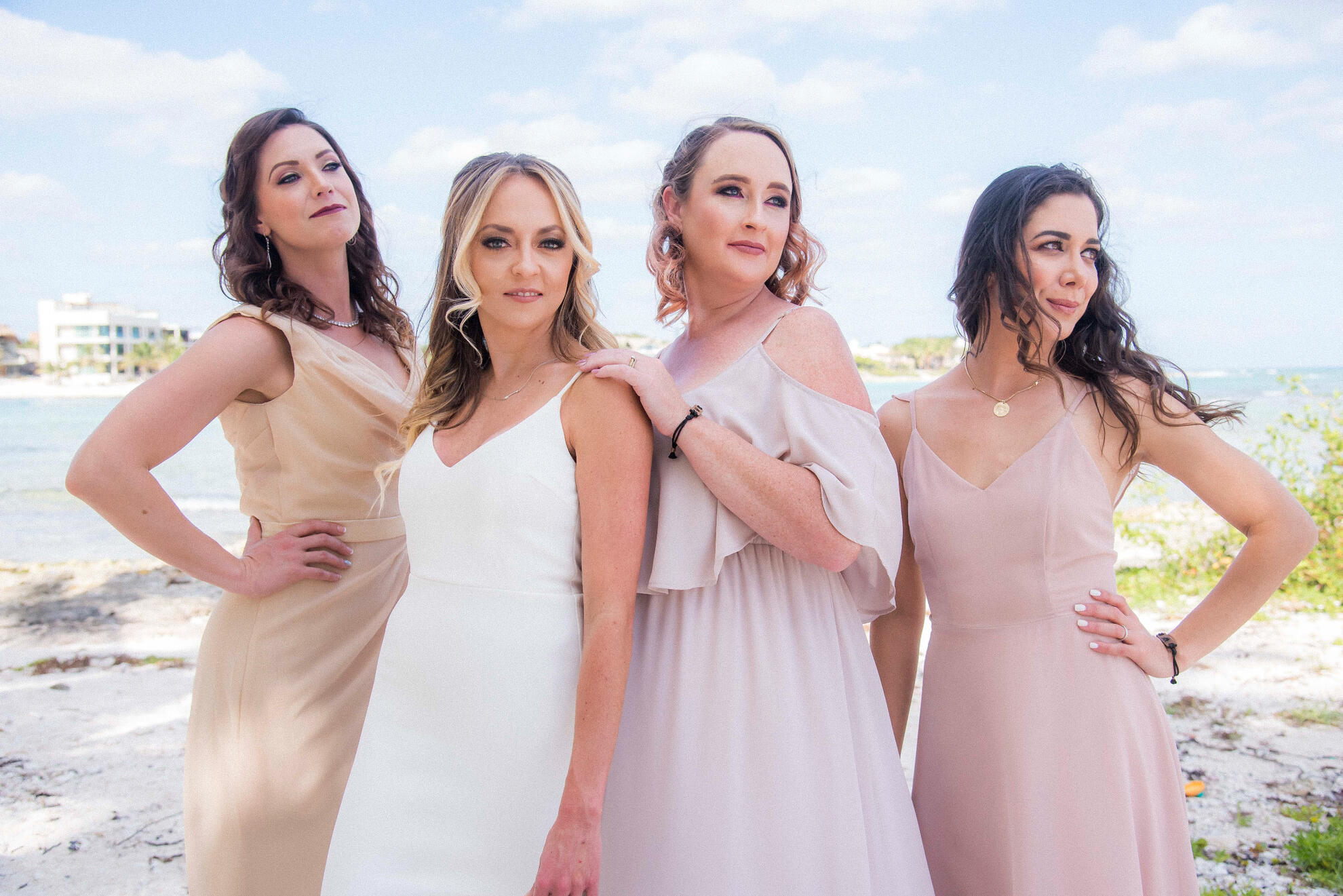 Bride surrounded by three women in soft pastel gowns, standing on coastal terrain under a cloudy sky — captured with warmth and editorial flair, reflecting grounded feminine presence.