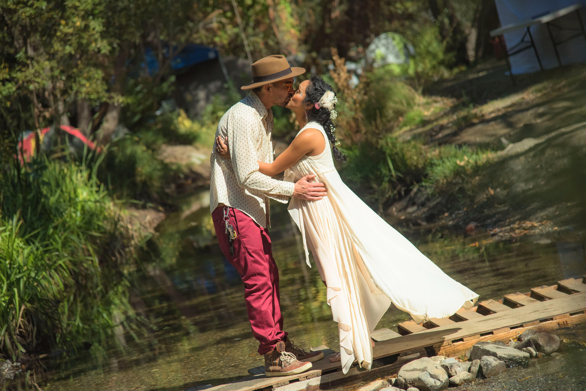 Couple kissing barefoot on a wooden bridge in the forest, framed by soft sun and greenery — intimate, grounded, and free-spirited wedding portrait in nature.