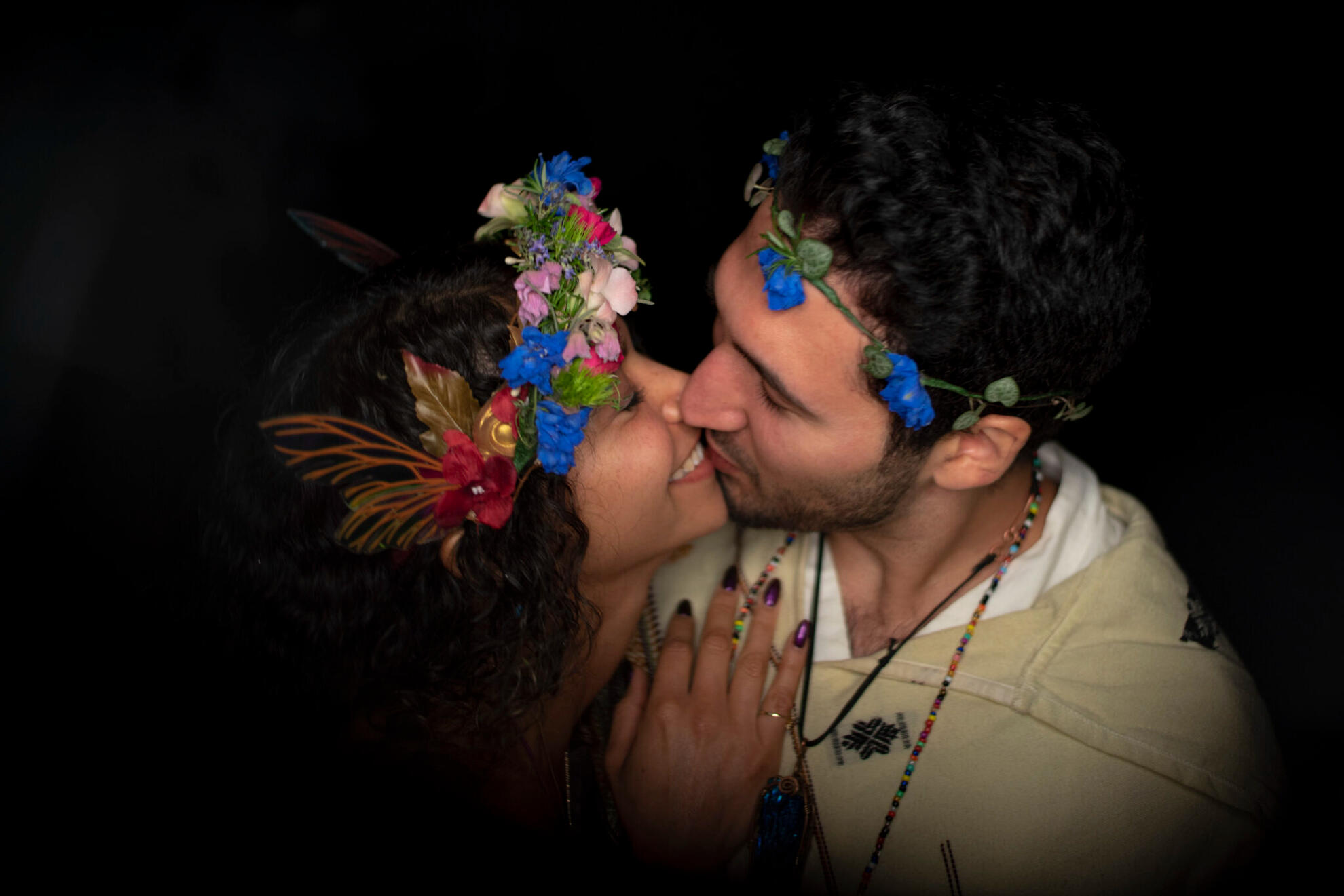Couple in a ceremonial wedding moment, adorned in flower crowns, sharing an intimate kiss under low light — captured in a documentary style emphasizing presence and emotion.