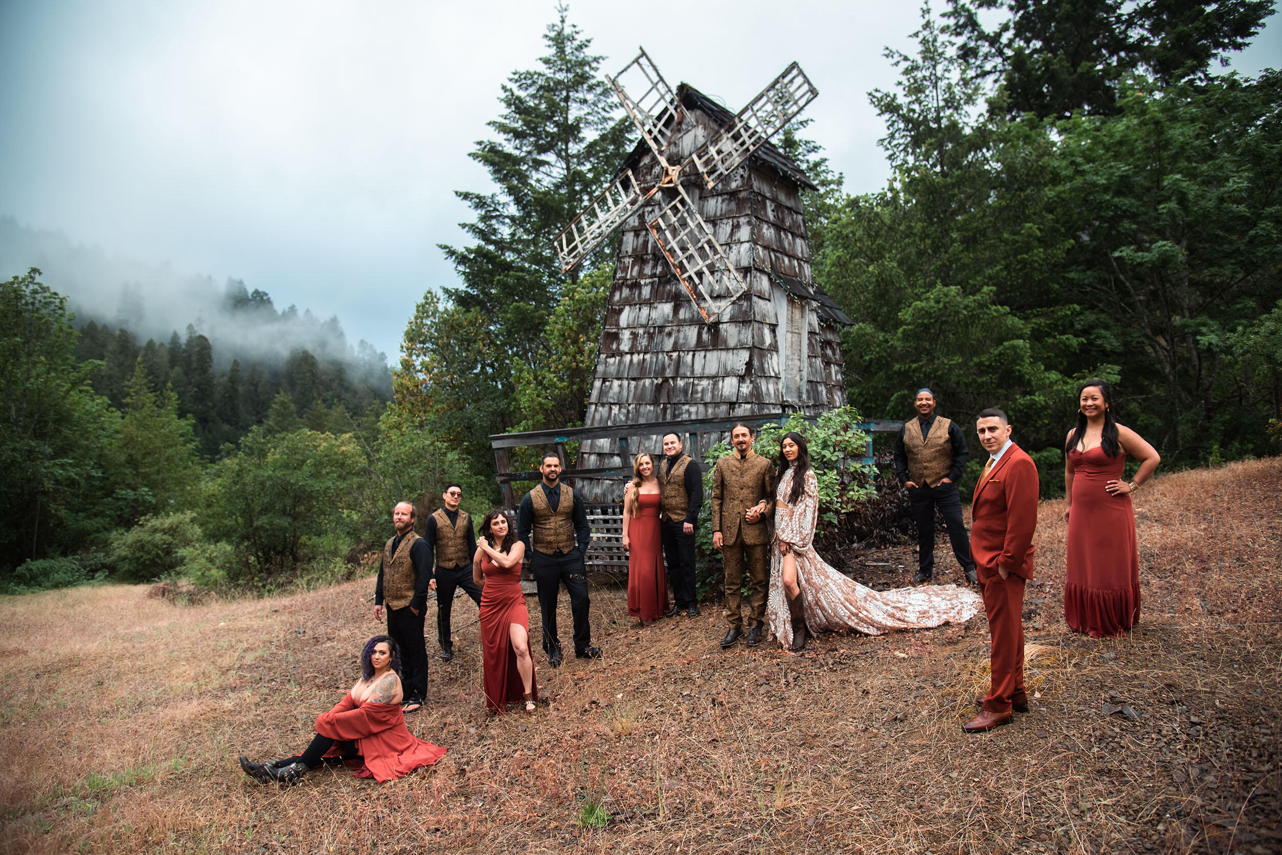 Wedding party dressed in earthy reds and browns posed in front of a rustic windmill, surrounded by forest and fog — dramatic, grounded, and mythic atmosphere.